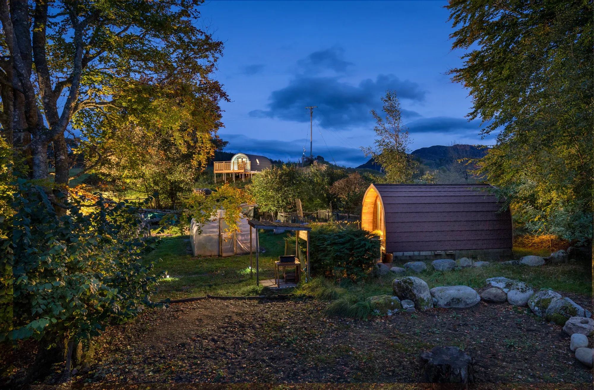 Cosy Coorie Pod glowing at dusk among autumn trees, with Muckle Meadow Pod lit on the hillside behind