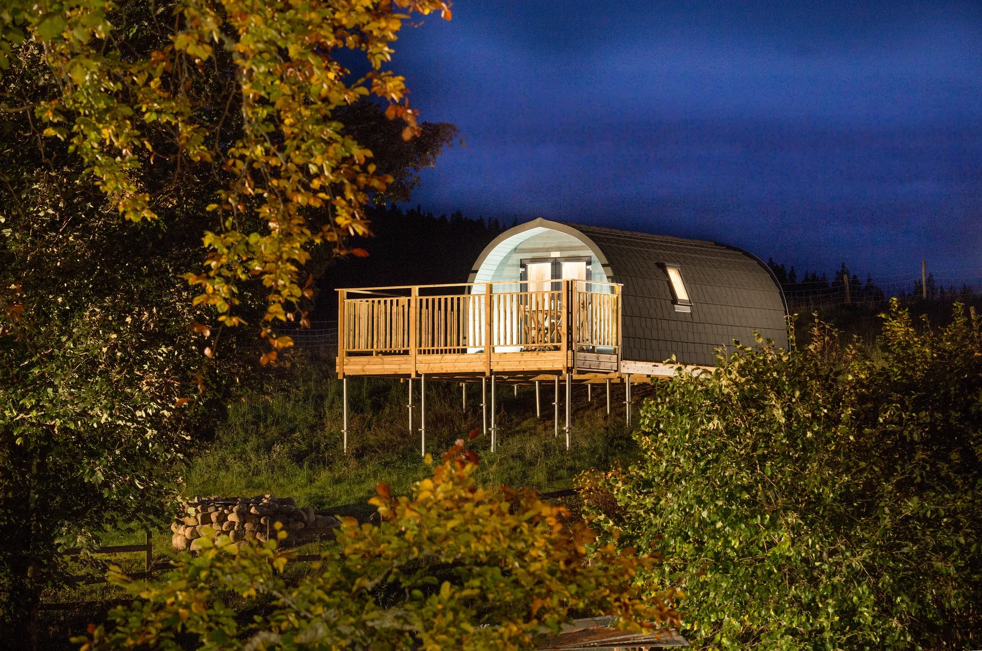 Muckle Meadow Pod at dusk — lit deck glowing against a deep blue sky, framed by autumn trees
